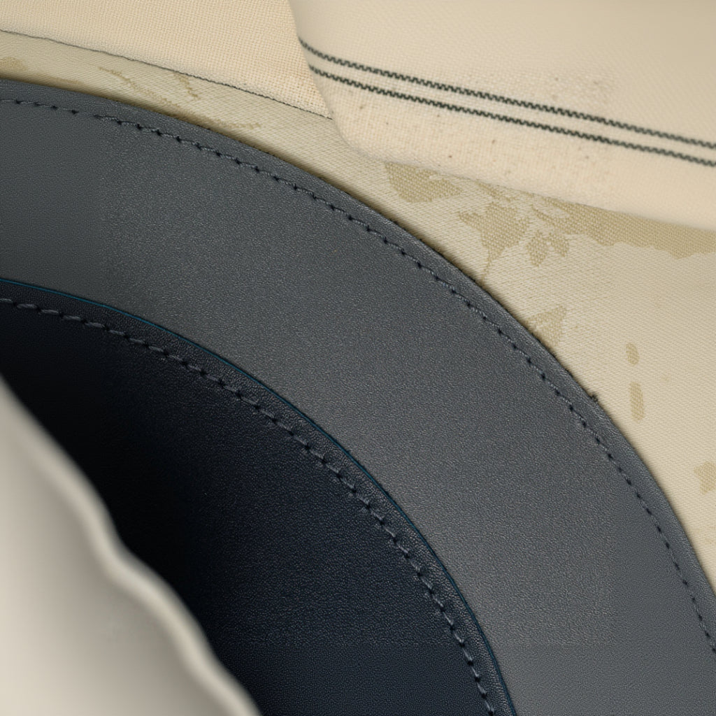 A high-angle close-up highlighting the intricate construction of a dual-textured leather placemat. The image shows the curved border with neat, double-row perimeter stitching that seamlessly joins two different leather finishes. The placemat is positioned over a light-colored floral tablecloth and a striped fabric napkin.