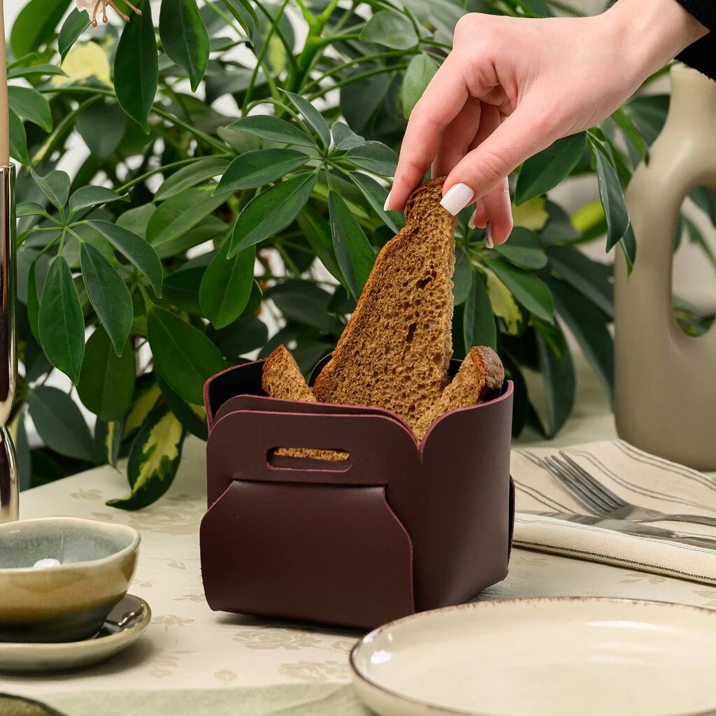 A hand with white manicure placing a slice of dark rye bread into a burgundy leather bread basket