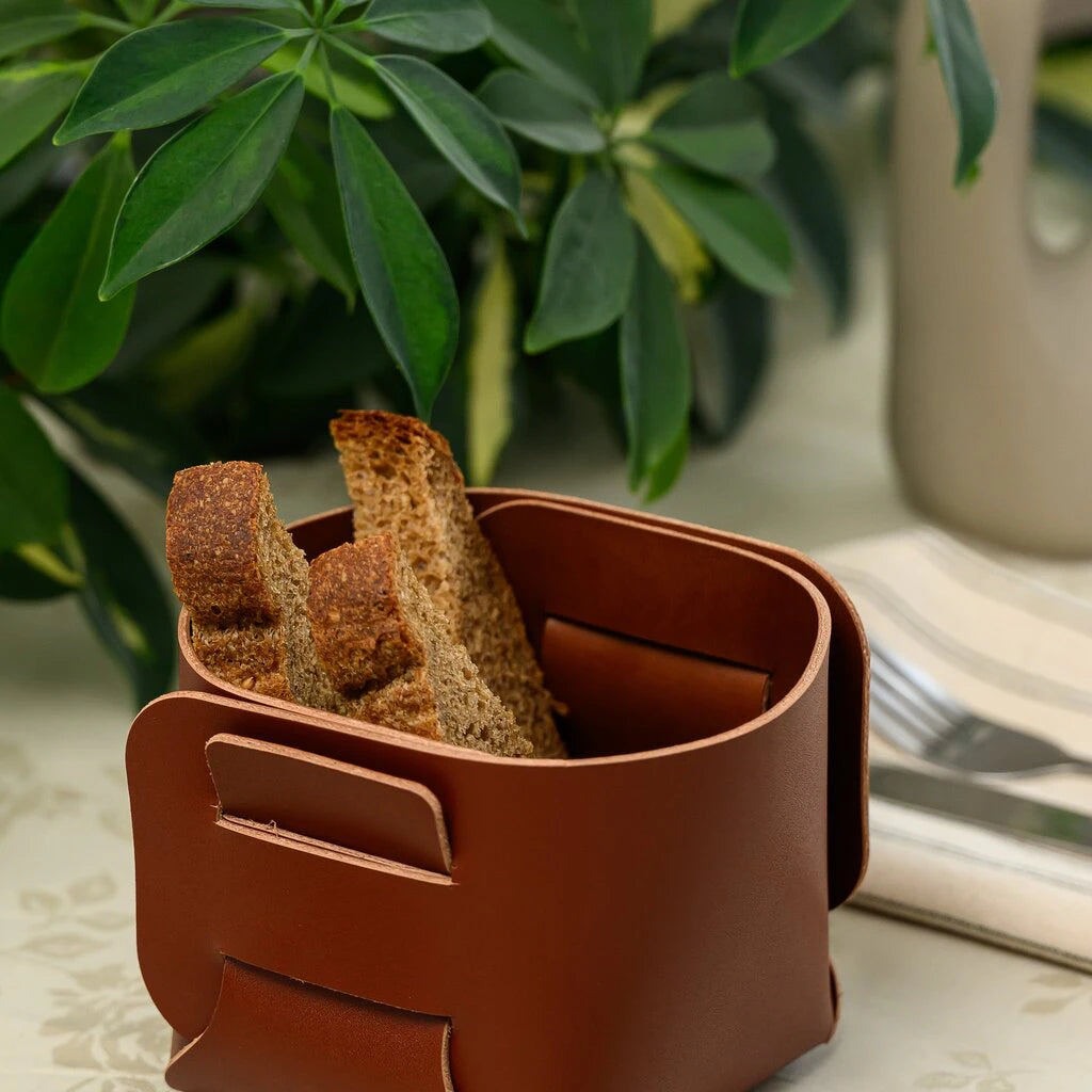 Square leather bread basket with rye bread slices placed on a table with a green plant background