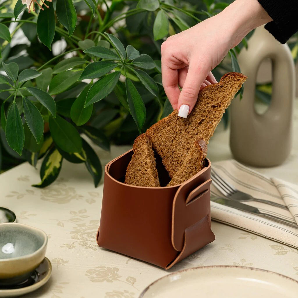 Elegant serving suggestion featuring a handcrafted leather bread basket. A warm, homey scene showing the practical use of this minimalist tabletop accessory with fresh dark bread.
