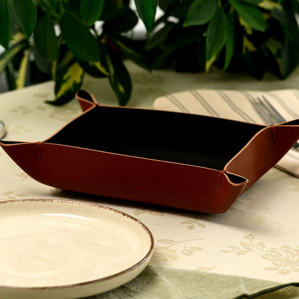 A premium rectangular placemat made from high-quality brown genuine leather with a smooth matte finish. The image shows a top-down view of the placemat on a light wooden table, styled with a ceramic plate, cutlery, and a linen napkin. It highlights the durable material and elegant minimalist design for upscale restaurant table settings