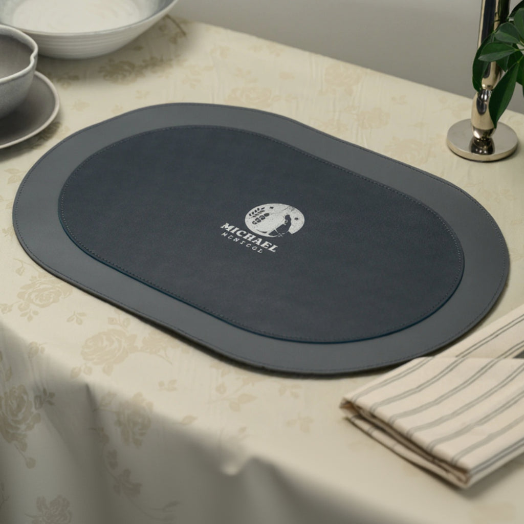 An oval-shaped leather placemat with a dual-textured design, featuring a smooth outer border and a napped inner inlay. The two sections are joined by precise decorative stitching. A circular logo with the name "MICHAEL MCNICOL" is embossed in the center. The placemat is presented on a floral-patterned tablecloth as part of a classic table setting