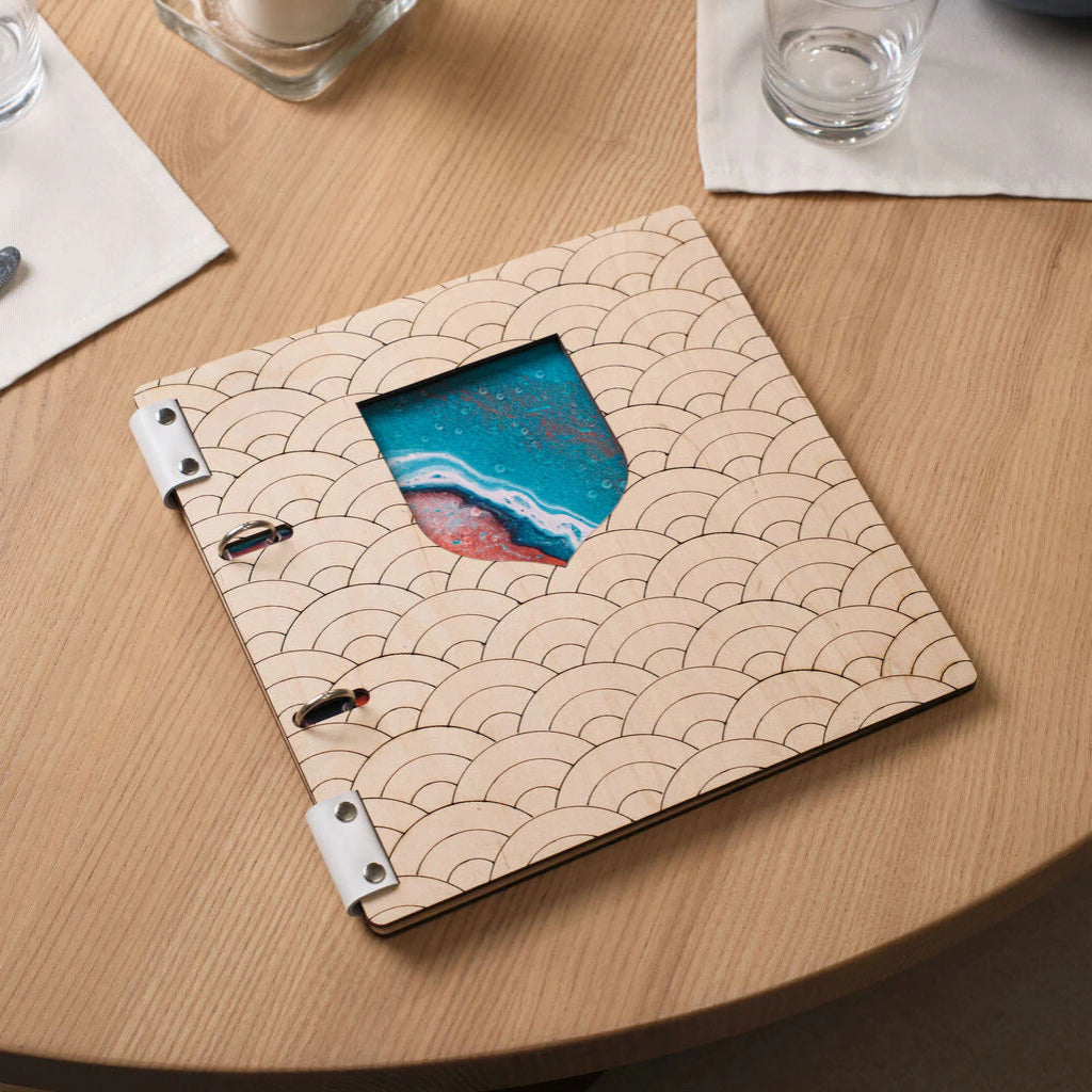 A high-angle lifestyle shot of a premium custom menu folder featuring a distressed blue wooden cover with a white topographic map line pattern. The menu is held together by black genuine leather straps and brass rivets. It is placed on a light oak wooden table in a sophisticated restaurant setting, surrounded by a navy blue ceramic plate, a clear water glass, and a wine glass, emphasizing a luxury dining experience.