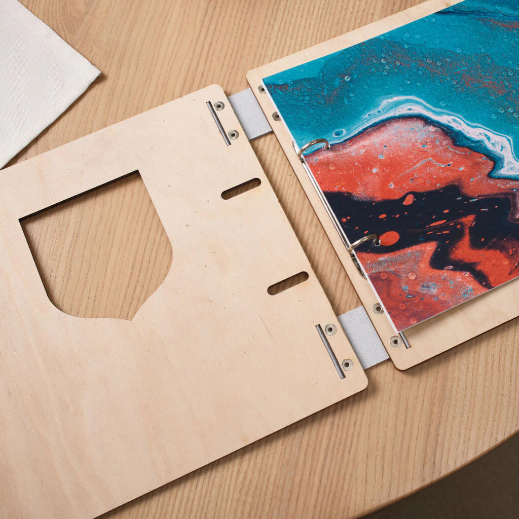A professional lifestyle photograph of a bespoke blue wooden menu cover laid flat on a light-colored oak dining table. The cover features an intricate, laser-engraved white topographic map pattern that covers the entire surface. A unique semi-circular window cutout in the center reveals a vibrant cocktail image inside. The menu is accented with premium black leather hinges and polished brass rivets. I