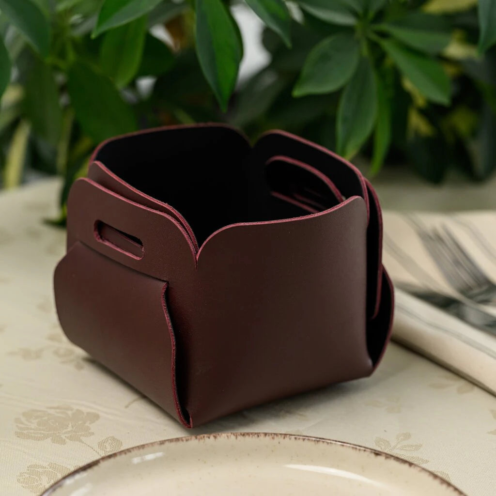 A rectangular brown leather bread basket with corners secured by tied leather laces, sitting on a floral tablecloth