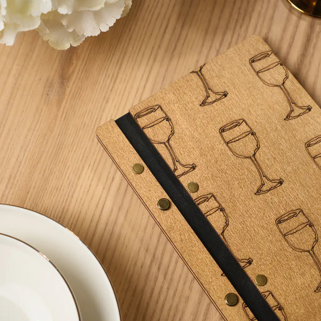 A detailed close-up of the menu folder's hinge area, displaying the contrast between the natural oak wood, the black leather-like spine strip, and the gold-toned metal fasteners.