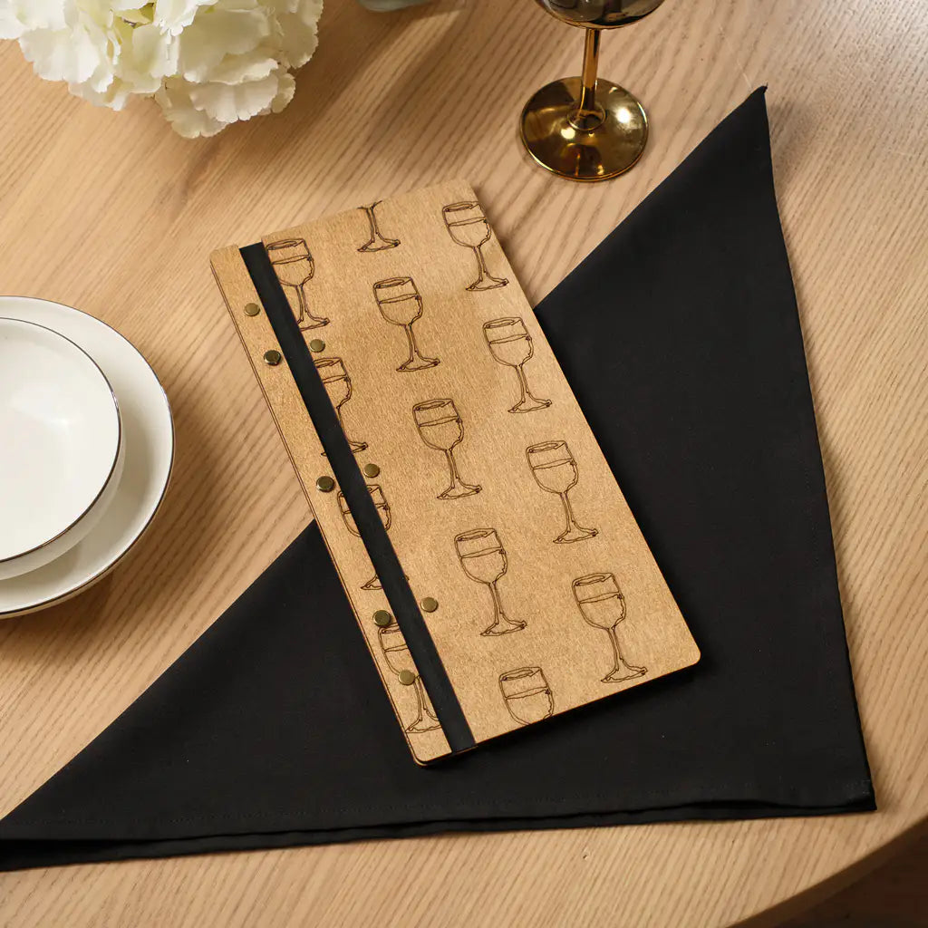 A top-down view of the closed wooden menu folder resting on a black triangular-folded napkin, highlighting the repetitive engraved wine glass motif and the black spine.
