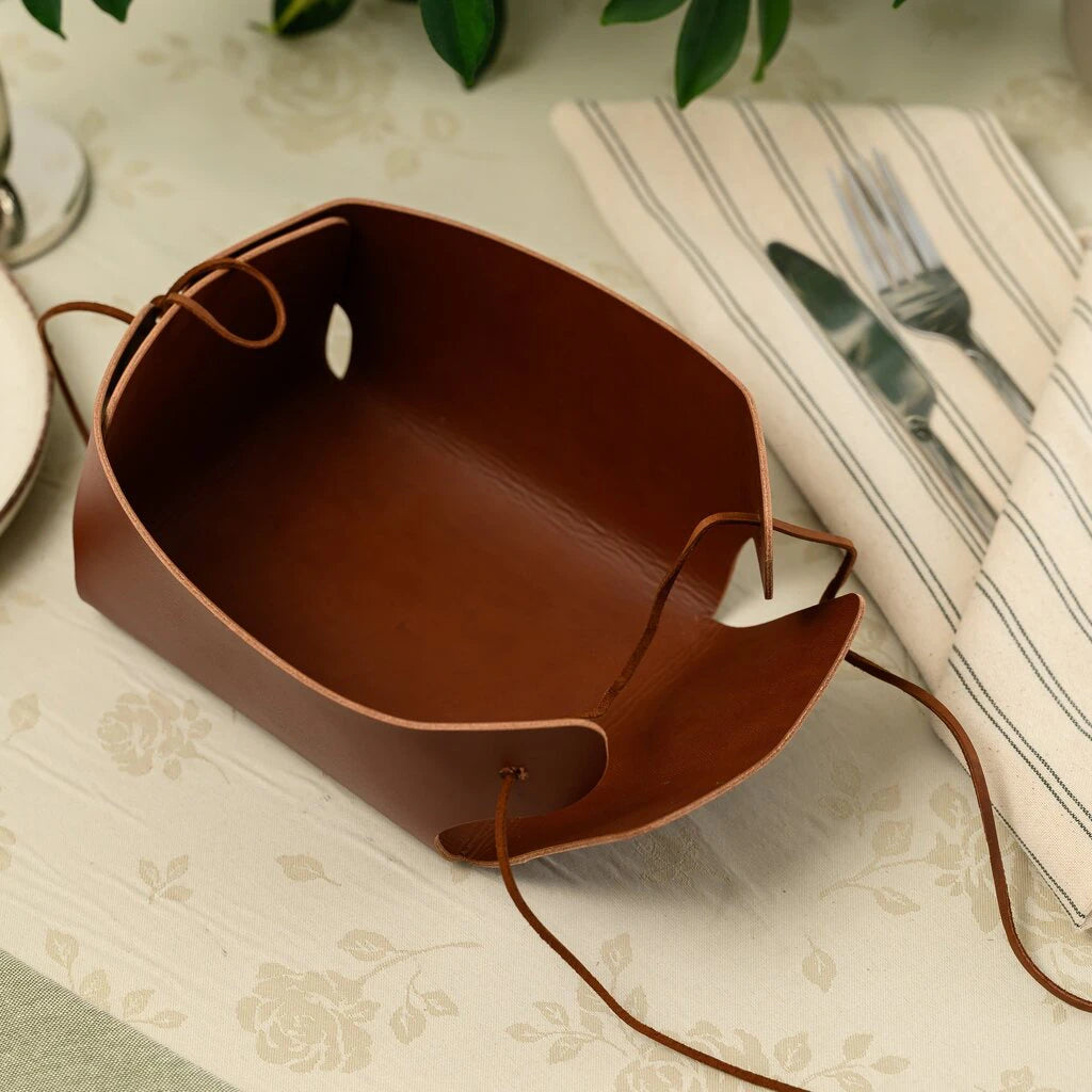 Demonstration of the foldable design of a brown leather bread basket with long leather ties, showing how the flat leather sheet transforms into a functional serving tray