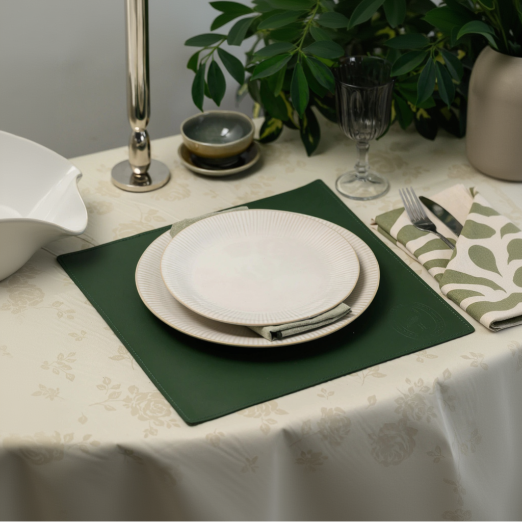 A top-down lifestyle shot of a square dark green leather placemat on a cream floral tablecloth, set with white ceramic plates, a patterned napkin with cutlery, and elegant glassware.