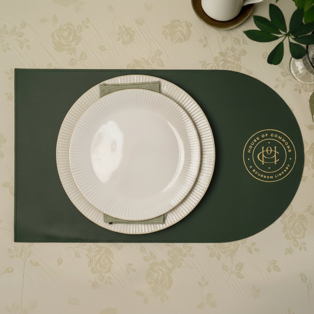 Dark green leather placemat with rounded silhouette, crafted with a smooth premium finish and detailed with gold foil embossing, styled in an elegant table setting with white ceramic plates and linen napkin on a soft floral tablecloth, refined horeca table decor, ideal for fine dining restaurants, cafes, weddings, and sophisticated hospitality interiors