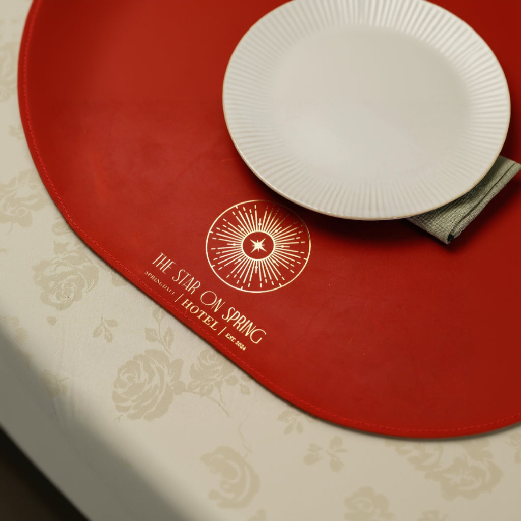 Luxury round red leather placemat with premium gold foil logo 'THE STAR ON SPRING HOTEL', featuring a white ribbed plate and sage green napkin on a floral tablecloth
