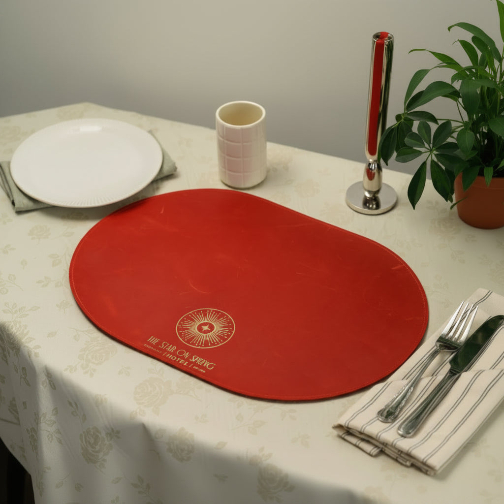 Wide shot of an oval red leather placemat with a gold foil hotel logo, styled on a floral tablecloth with silver cutlery, a white cup, and a green houseplant background