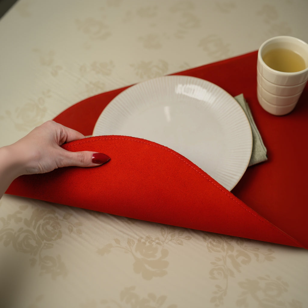 A hand lifting the edge of a flexible oval red leather placemat to demonstrate its premium quality and soft suede-like underside