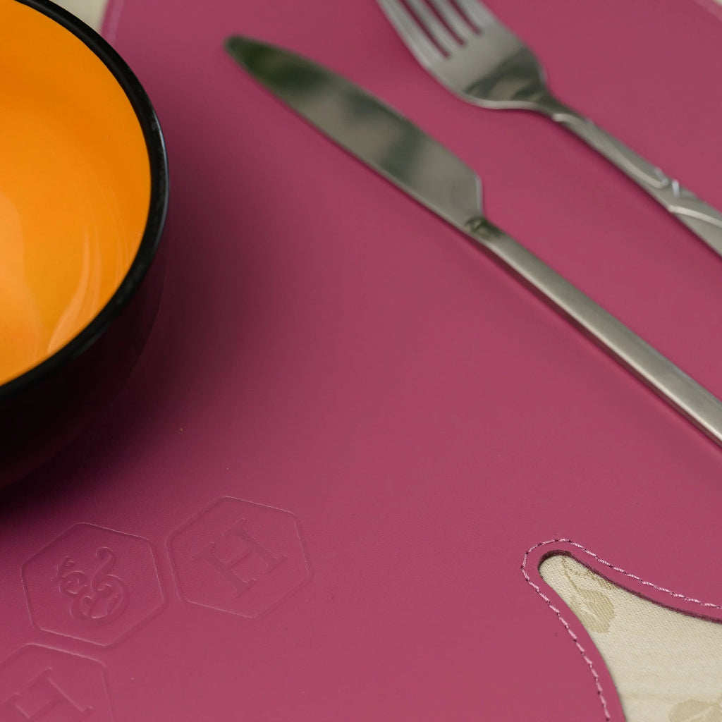 A vibrant magenta leather placemat featuring an embossed hexagonal logo. A stainless steel knife and fork are placed neatly on its surface next to a black bowl with a bright orange interior.