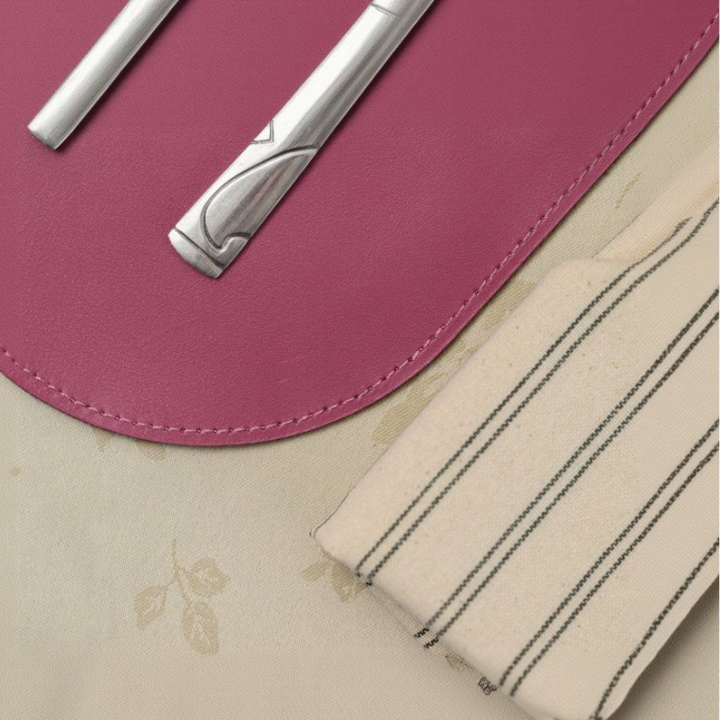 Top-down close-up of a rounded magenta leather placemat with neat edge stitching. Silver cutlery rests on the placemat, which is laid over a floral tablecloth next to a striped linen napkin.