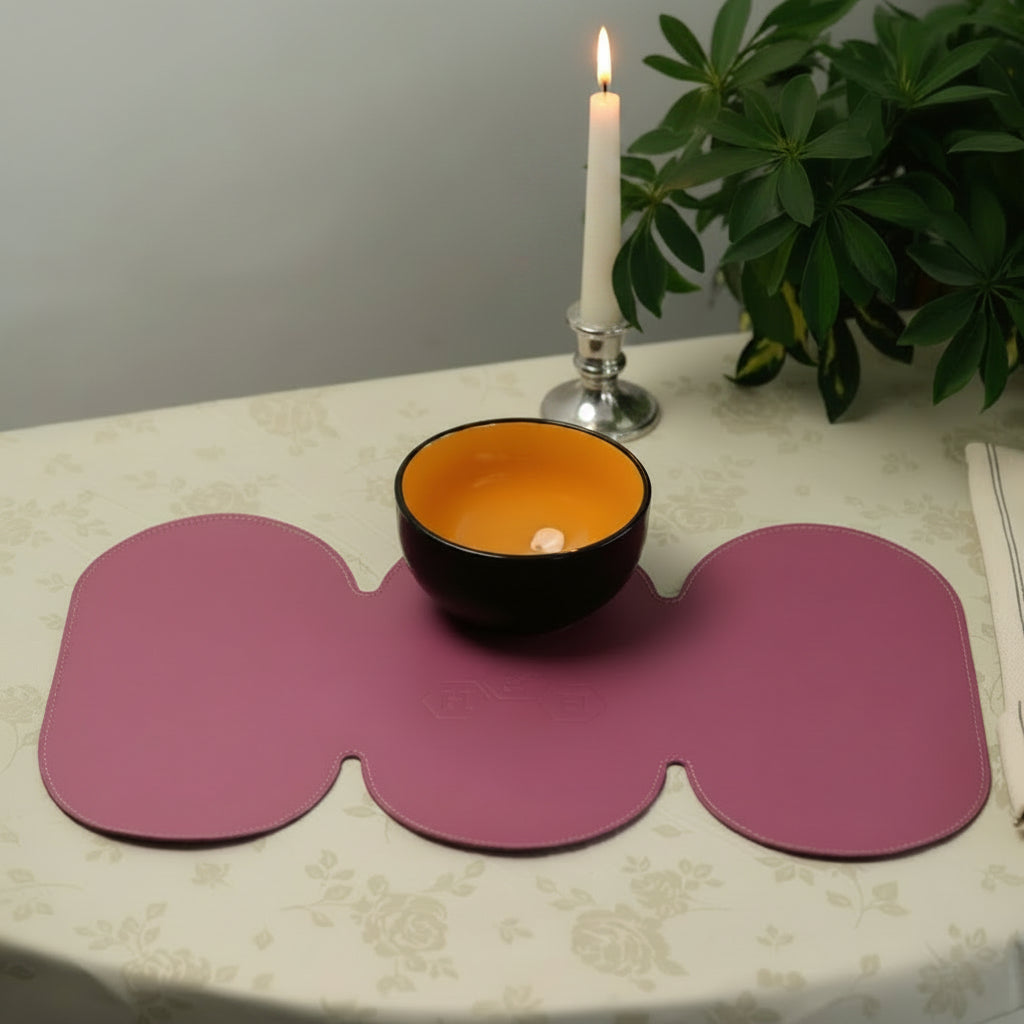 A top-down lifestyle photograph of a luxurious magenta pink leather table runner. The runner has a distinctive scalloped, triple-oval shape and features precise perimeter stitching and subtle debossed detailing. It is styled on a cream floral tablecloth with a black and orange bowl, a burning silver candlestick, and green foliage