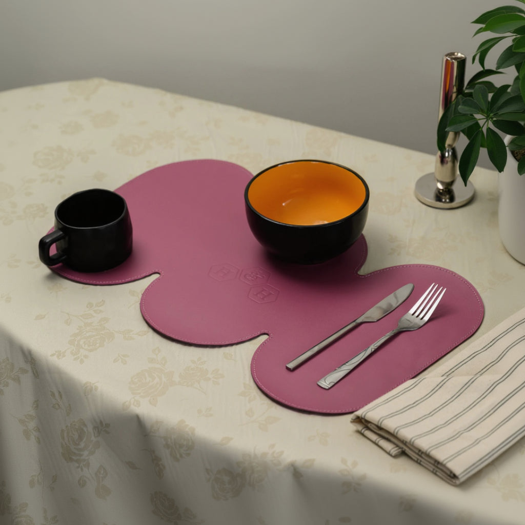 A uniquely shaped, wavy magenta leather placemat set on a floral tablecloth. It holds a black mug, an orange-lined bowl, and silver cutlery, showcasing its large size and triple-lobed design.