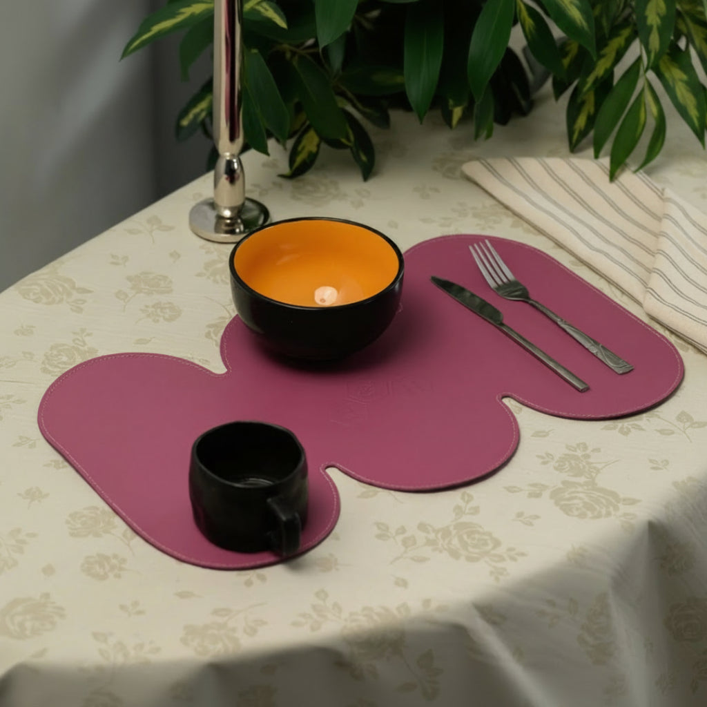 A long, uniquely shaped magenta leather placemat styled on a cream floral tablecloth. The placemat features a modern black mug, a bowl with a bright orange interior, and silver cutlery, creating a stylish table setting.