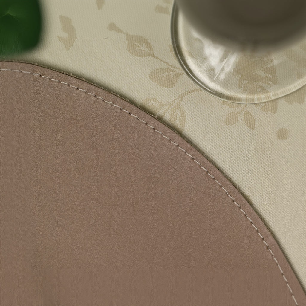 An extreme close-up of a mocha-colored leather placemat, focusing on the high-quality craftsmanship. The shot highlights the neat, light-colored decorative stitching along the rounded edge and the smooth, fine-grained texture of the leather. In the blurred background, a cream floral tablecloth and a striped fabric napkin provide a soft contrast, emphasizing the placemat's elegant and durable finish