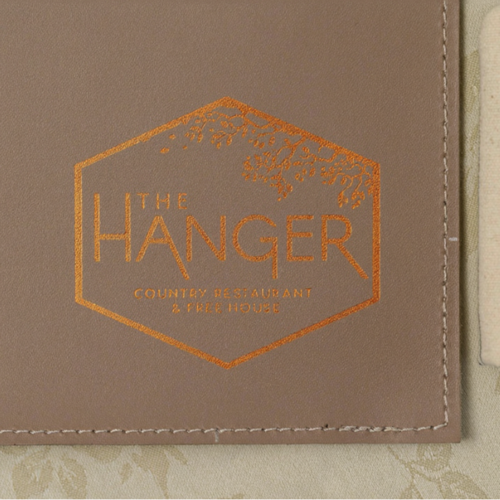 A macro shot of a leather placemat featuring a detailed copper embossed logo in the corner. The logo is enclosed in a hexagon with a tree design and the text "THE HANGER COUNTRY RESTAURANT & FREE HOUSE." The image highlights the fine, grainy texture of the leather and the precise stitching along the straight edge. A striped fabric and floral tablecloth are visible in the background, emphasizing the professional and branded look of the product.