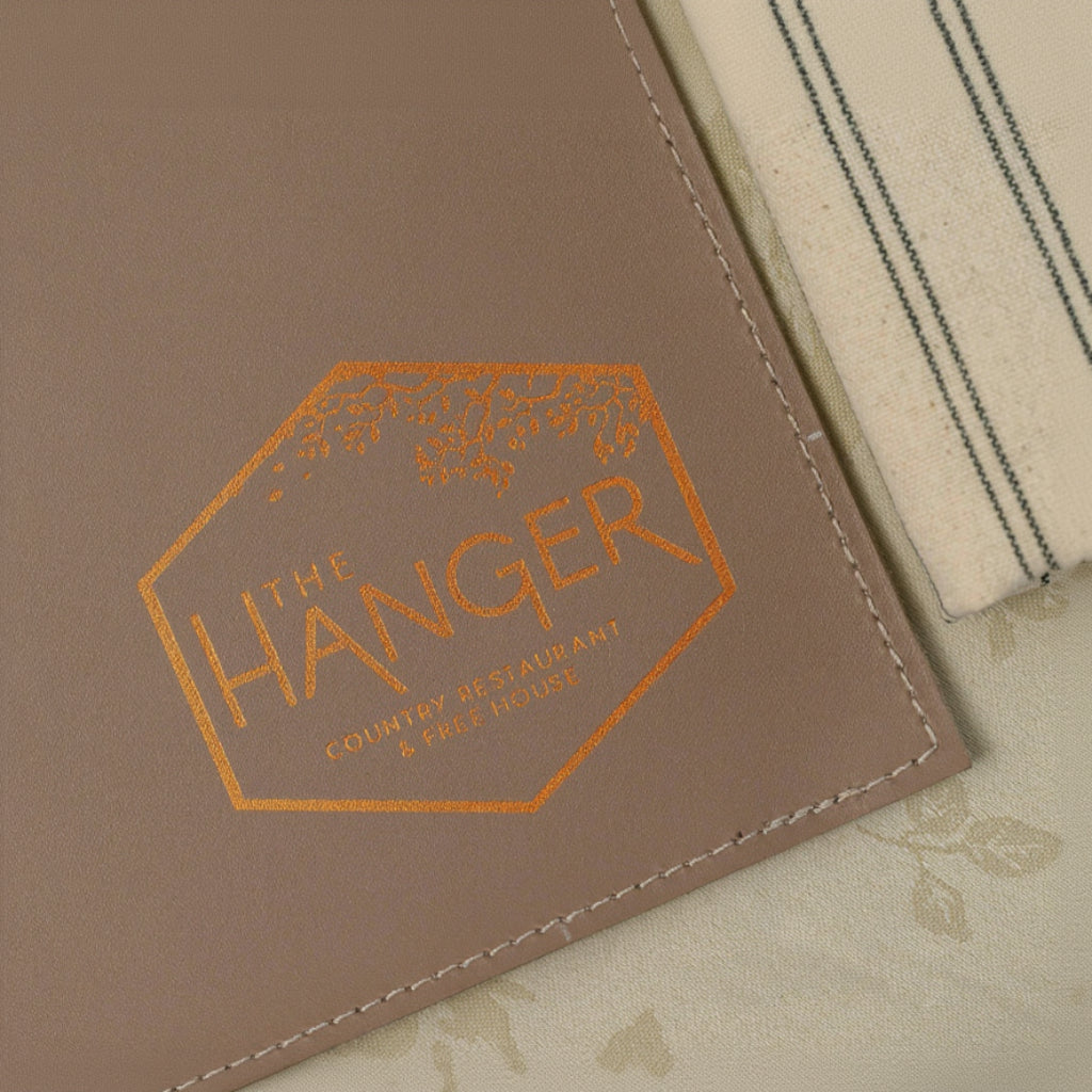 A close-up, top-down view of a leather placemat featuring visible stitching along the edges. A metallic hexagonal logo is embossed on the surface, containing the text "THE HANGER COUNTRY RESTAURANT & FREE HOUSE" topped with a delicate branch and leaf design. The placemat is partially layered over textured, patterned fabric napkins.