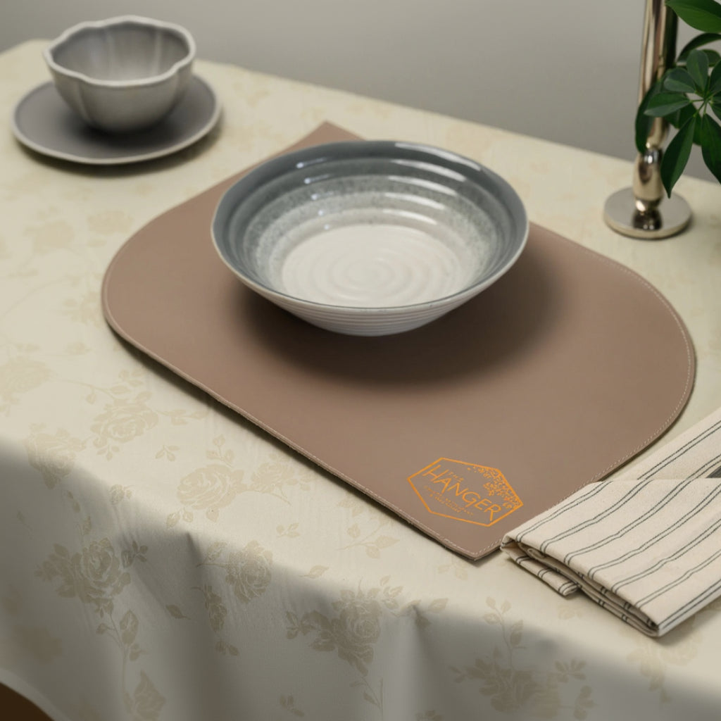 A full view of a table setting featuring a leather placemat with a unique shape—rectangular with two diagonally rounded corners and visible perimeter stitching. A metallic hexagonal logo for "The Hanger" is embossed in the lower corner. On the placemat sits a ceramic bowl, while a striped fabric napkin with cutlery, a glass, and a small side dish are arranged around it on a floral-patterned tablecloth.