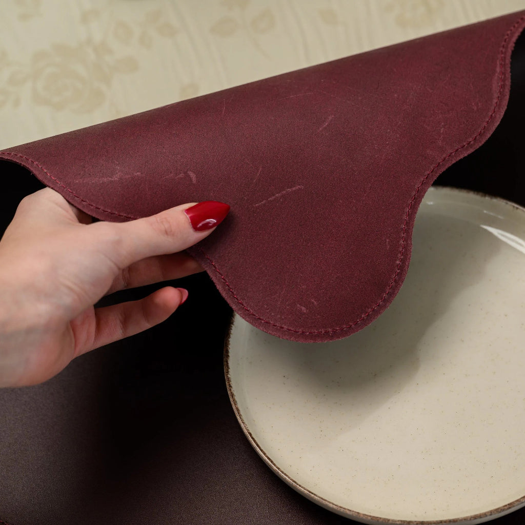 A close-up shot of a hand lifting a premium burgundy leather placemat with a scalloped edge. The image highlights the natural matte texture of the leather and detailed perimeter stitching next to a cream ceramic plate