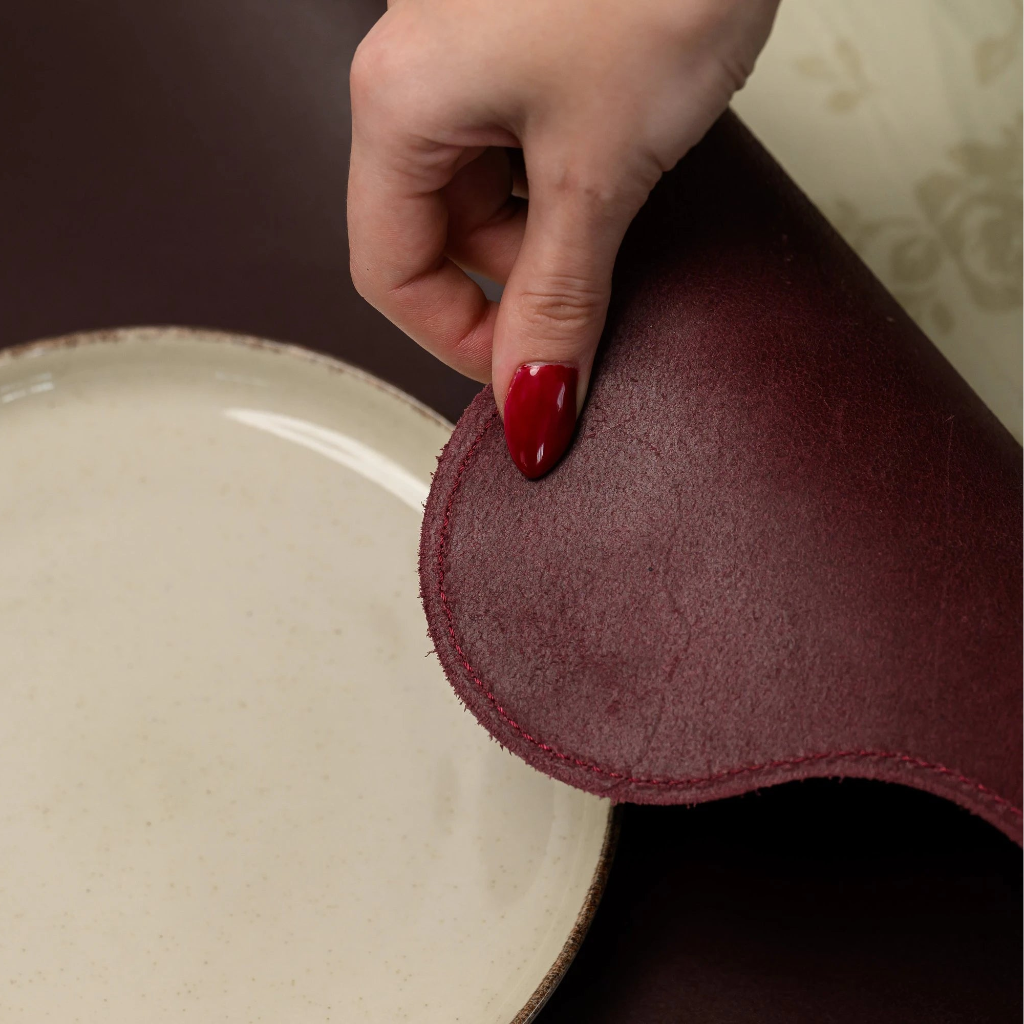 A close-up showing a hand lifting the edge of a flexible burgundy leather placemat to reveal the natural soft underside and precision perimeter stitching. The mat is placed next to a cream-colored ceramic plate on a table with a floral tablecloth.