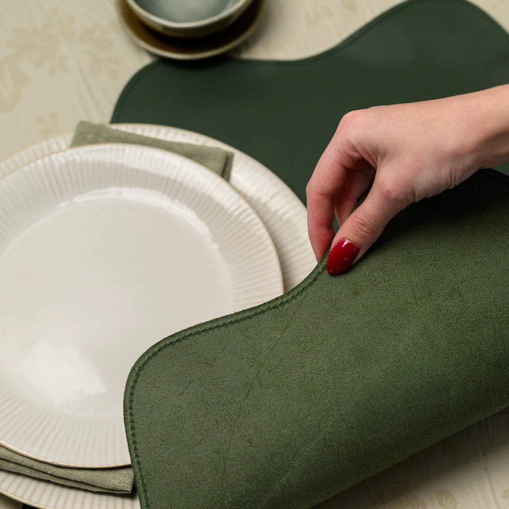 A close-up shot of a hand with red nails peeling back the edge of a forest green leather placemat to show its textured, light green suede backing. The focus is on the placemat's scalloped edge and the high-quality tonal stitching that follows its wavy contour. In the blurred background, white ribbed plates sit on a floral tablecloth, highlighting the placemat's role as a stylish and durable base for tableware
