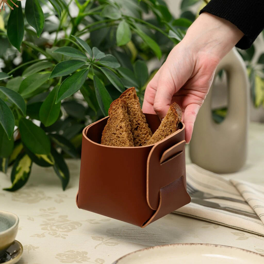 A person's hand is lifting a square leather bread basket filled with three slices of dark bread. The basket is held mid-air over a set dining table. In the background, there is a green houseplant and a modern beige vase, creating a cozy kitchen atmosphere.
