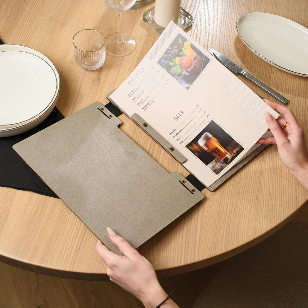A person's hands holding open a grey wooden menu folder on a restaurant table, revealing a beautifully designed interior page with sections for cocktails and beer, accompanied by vibrant beverage photography.