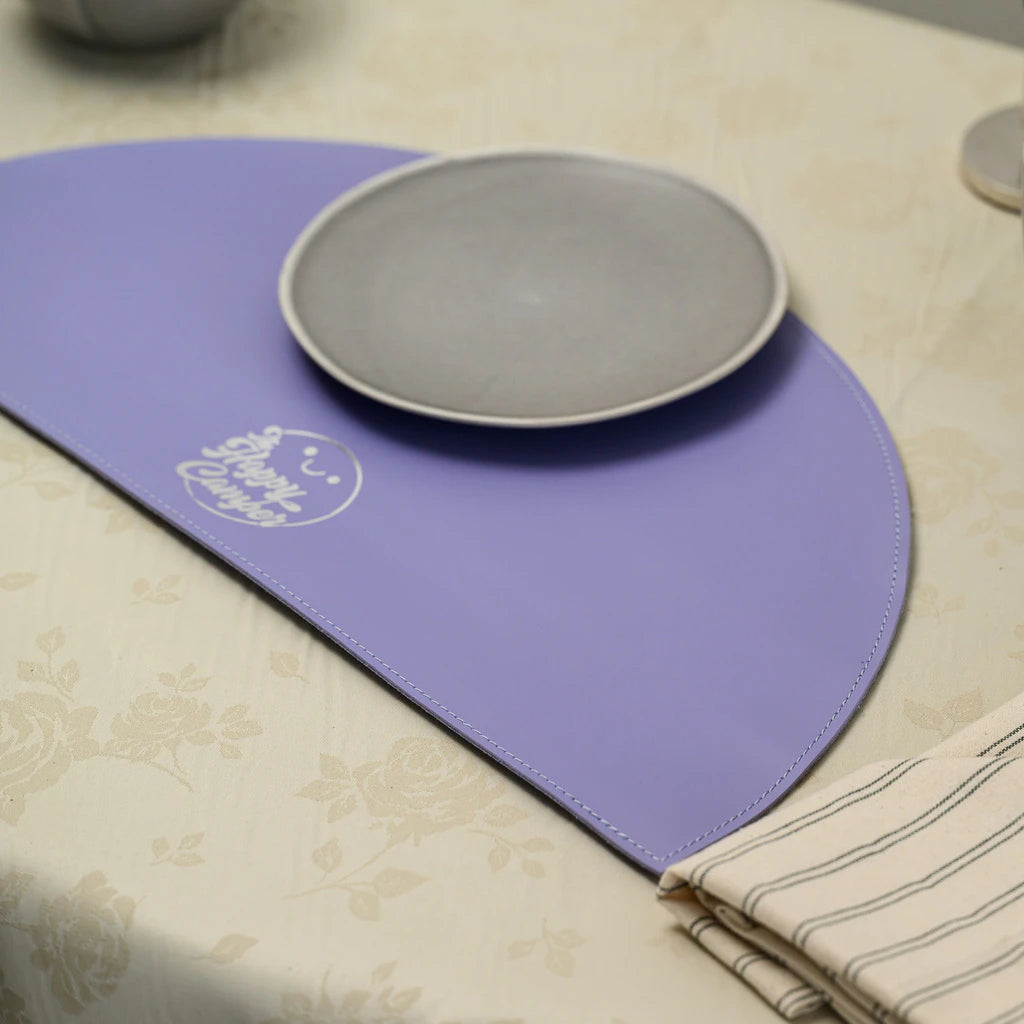 Macro shot of the silver-engraved "The Happy Camper" logo on the smooth lavender leather surface, with a grey plate visible in the corner