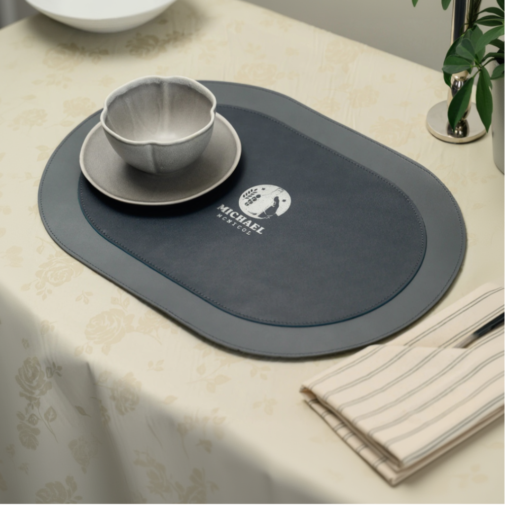 A top-down, slightly angled view of a dual-textured oval leather placemat. The design features a smooth outer border stitched to a textured, matte inner section. A circular personalized logo with the name "MICHAEL MCNICOL" is embossed in the center. The placemat is decorated with a ceramic bowl on a saucer and is set on a floral-patterned tablecloth next to a folded striped napkin.