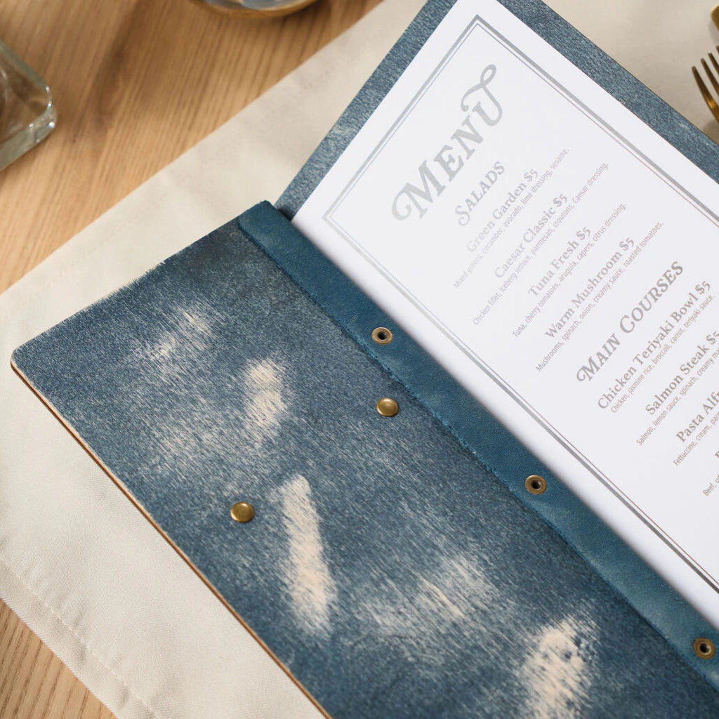 Close-up of the "jeans" blue wooden menu, highlighting the weathered texture and the blue leather spine strip secured with brass-colored rivets that hold the internal paper in place.