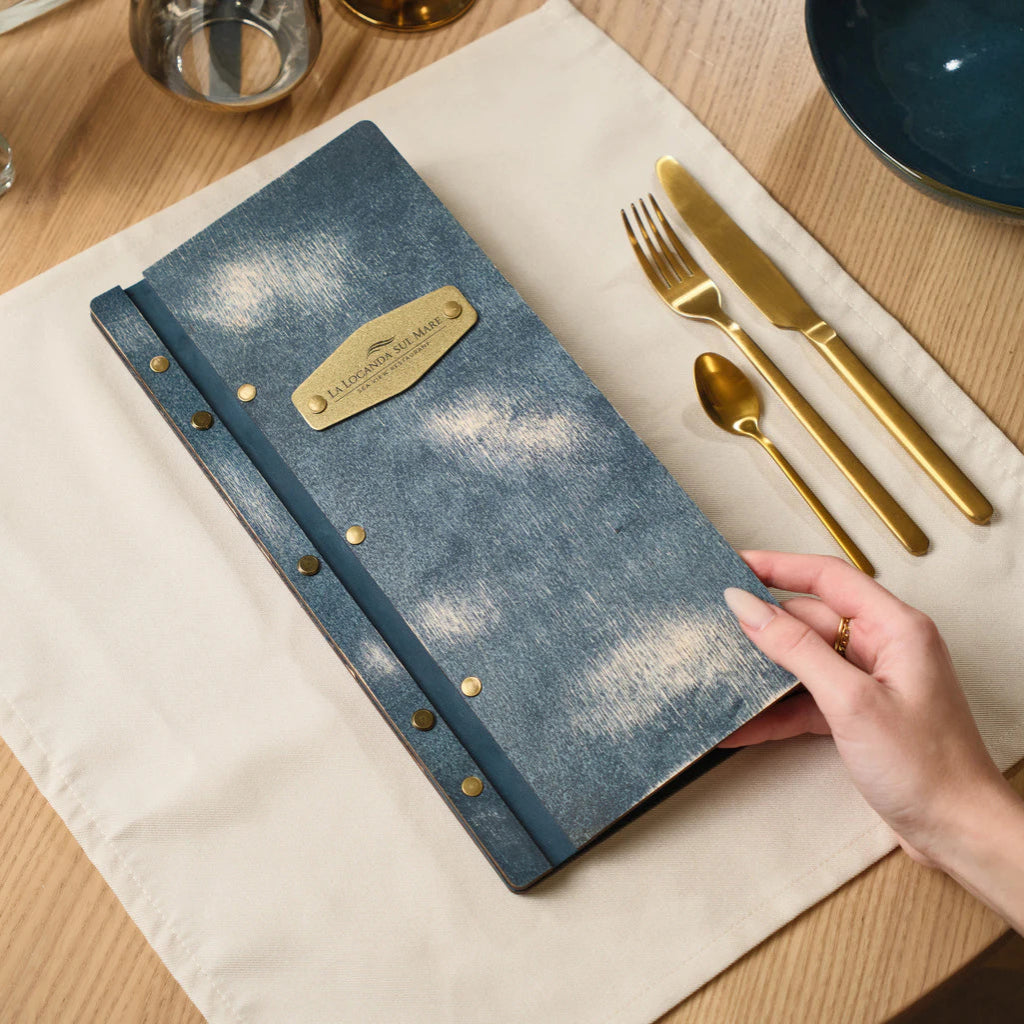 A person’s hand holding the blue wooden menu folder, revealing a brass plate on the cover engraved with the restaurant name "LA LOCANDA SUL MARE" and secured by small brass rivets.