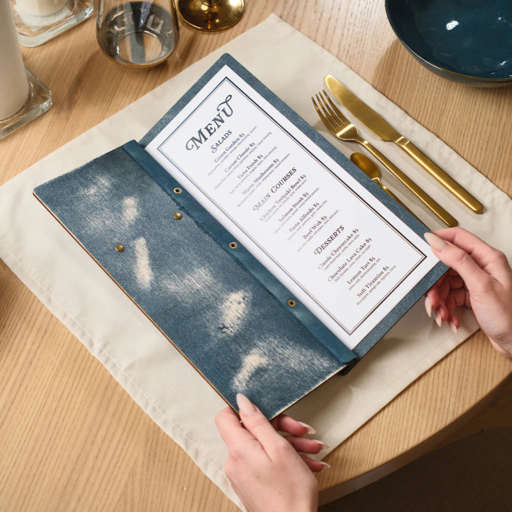 The blue distressed wood menu folder held open by two hands, showcasing a printed paper menu inside with categories for Salads, Main Courses, and Desserts.