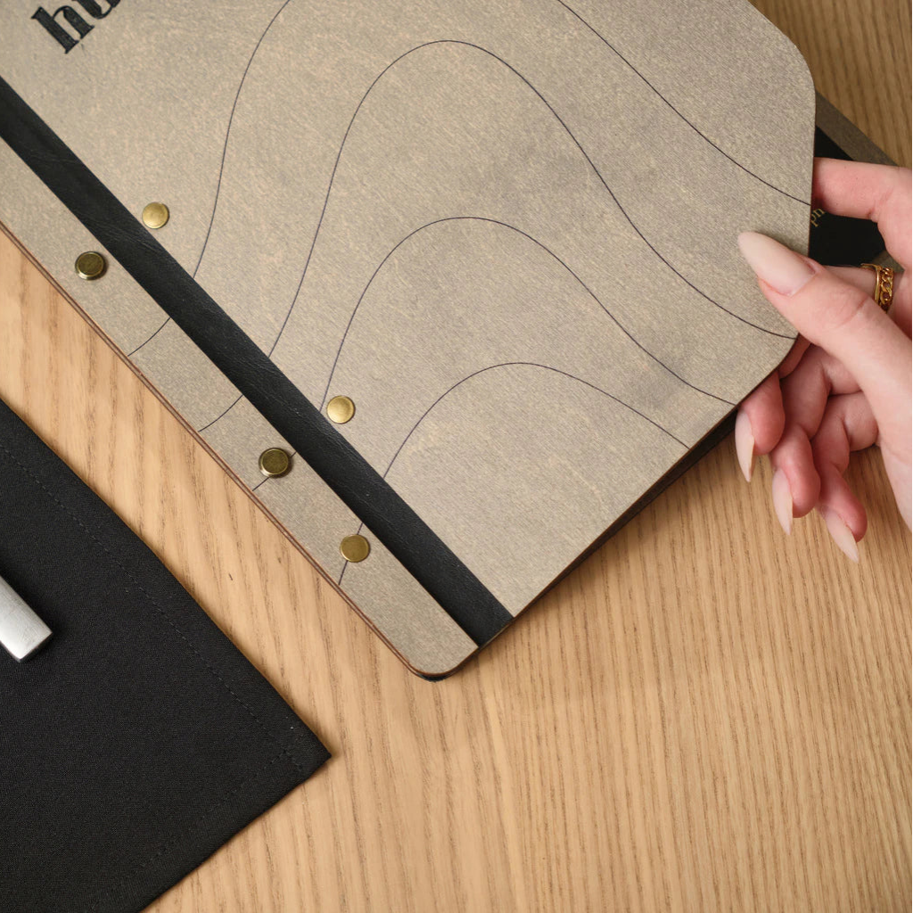 Close-up of a handcrafted wooden menu cover with a black leather spine and decorative gold studs, held by a hand to show the unique wavy engraving and texture