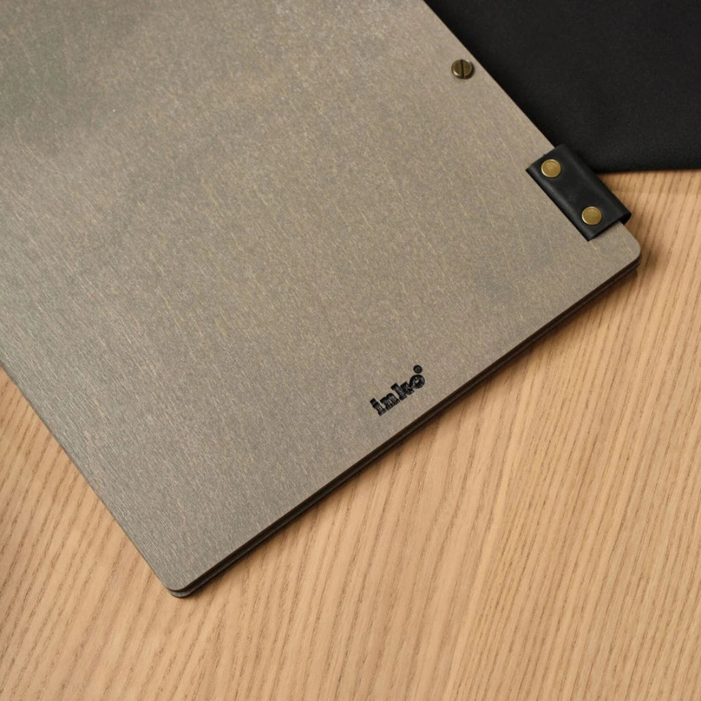 Custom grey wooden menu folder for restaurants featuring the engraved "inko" logo. This handcrafted wood menu cover includes flexible black leather strap accents and professional bronze bolt binding, presented on a modern wooden dining table
