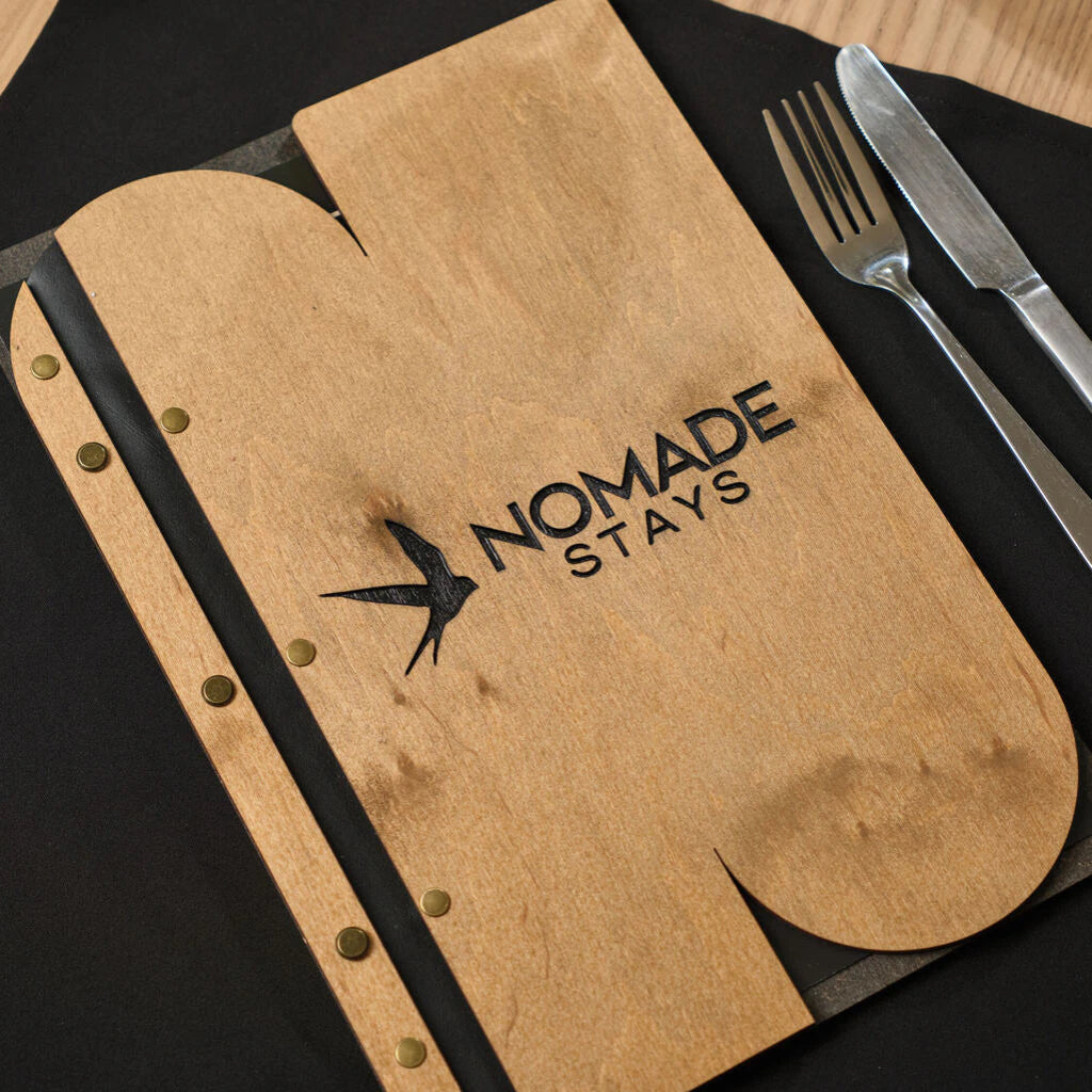 Wooden menu folder with a unique custom cutout and "NOMADE STAYS" logo engraving, styled on a table with a fork and knife