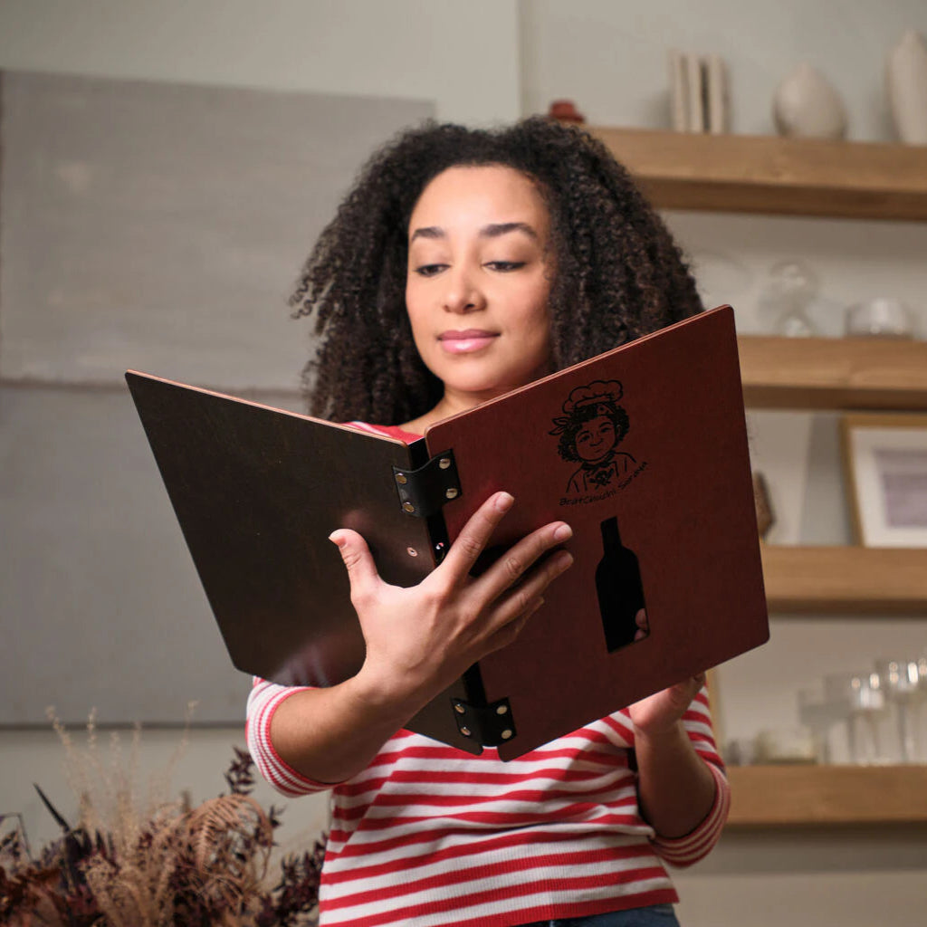 A woman holding open a chili-red wooden menu folder, revealing the dark wood interior and ring binder design in a modern, warm interior setting.