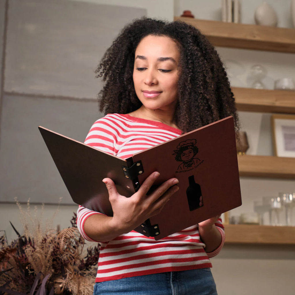 A woman with curly hair in a red striped shirt holds open a chili-red wooden menu binder, showing the dark wood interior and the engraved logo on the front.
