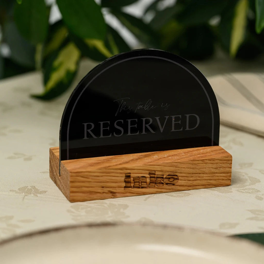 A close-up of a modern 'Reserved' table sign standing vertically on a light floral tablecloth. The sign is made of a solid wooden block with a glossy black acrylic face featuring the word 'RESERVED' in cutout letters. In the blurred background, a striped napkin, cutlery, and green plant leaves are visible