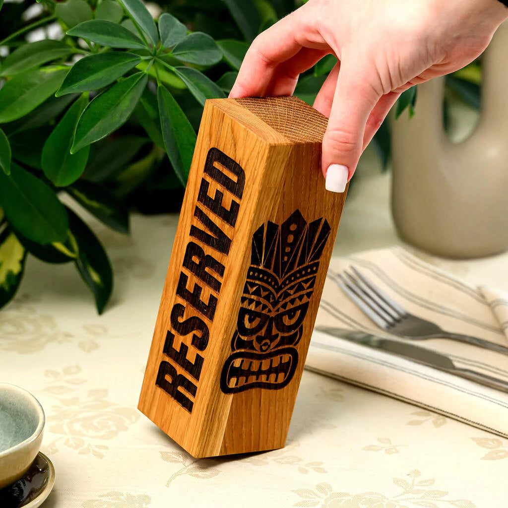 Handcrafted oak wood reservation sign with a tropical Tiki-themed engraving. Held by a hand in a restaurant setting, this durable and stylish table marker adds a premium touch to bar and cafe decor