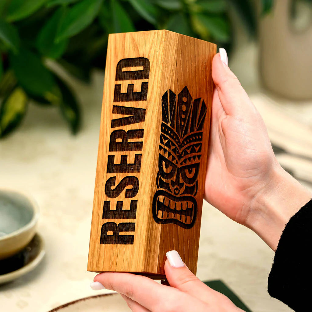 Detail view of a handcrafted oak reservation sign in a tropical Tiki-themed bar setting. Held between hands, the sign highlights the clean engraving and high-quality wood grain, adding an exotic and upscale feel to the table decor