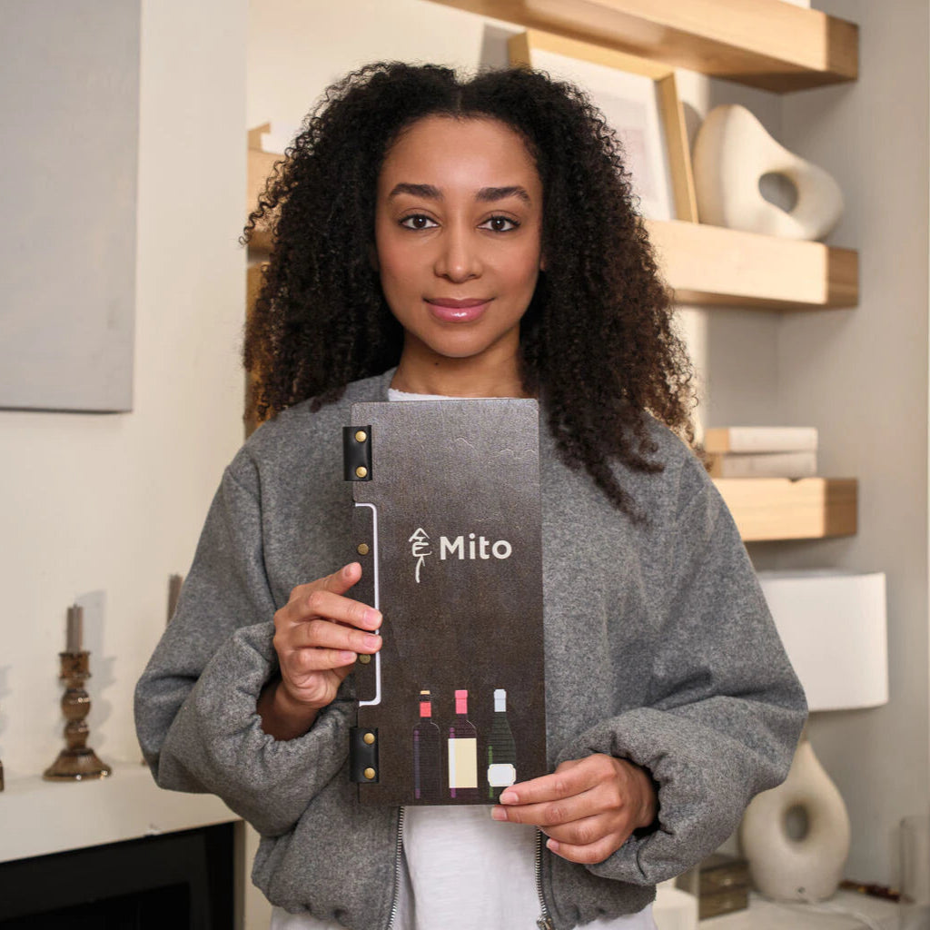 A woman holds the closed wenge menu folder vertically, providing a clear view of its slim proportions, the central "Mito" branding, and the decorative wine bottle illustrations.