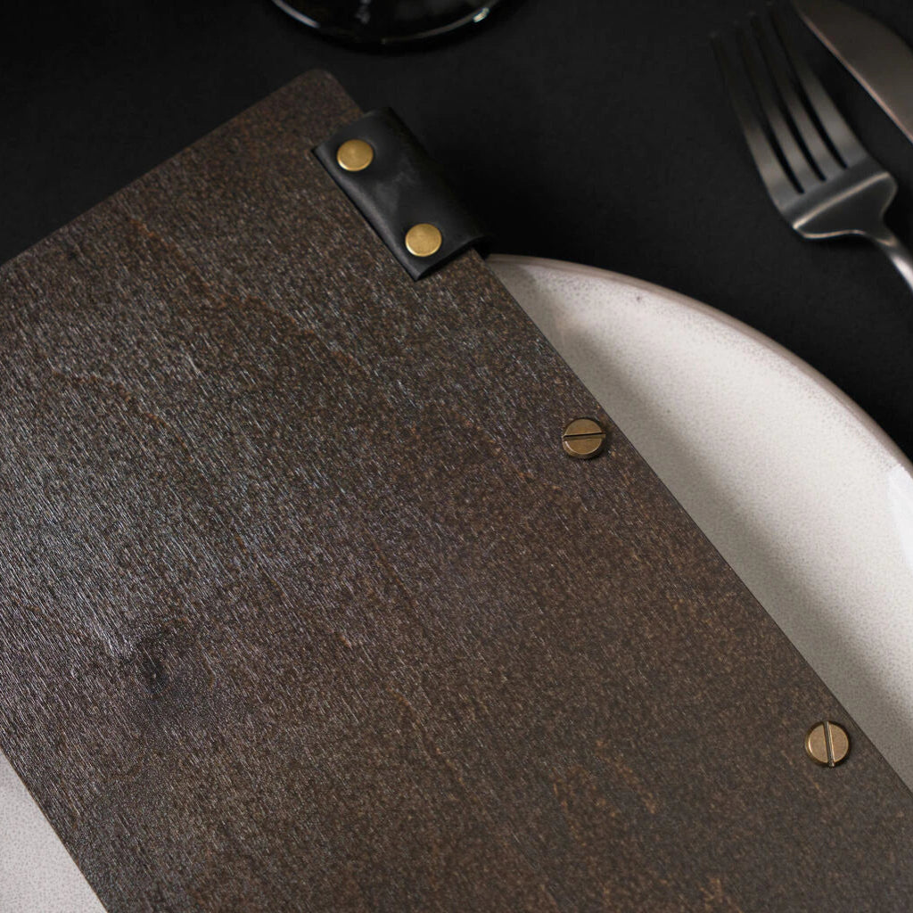 A top-down close-up of a closed dark wenge wooden menu folder resting on a white plate. The image highlights the brass-colored binding screws and a black leather hinge secured with brass rivets.