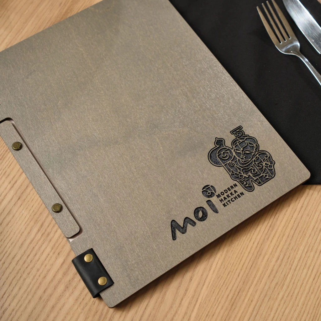 Close-up of a custom grey wooden menu folder for "Moi Modern Hakka Kitchen." Features professional laser-engraved branding, a unique black leather strap binding, and bronze metal hardware on a wooden table
