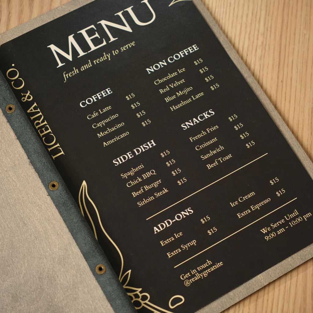 Angled close-up of a black restaurant menu card inside a gray wood-textured holder, showcasing elegant typography and gold-leaf style illustrations on the side
