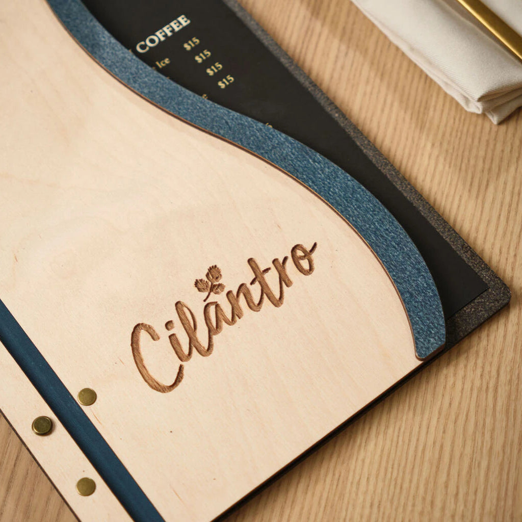 Macro close-up of a laser-engraved "Cilantro" logo on an ivory wooden menu cover, highlighting the textured blue wavy border and natural wood grain