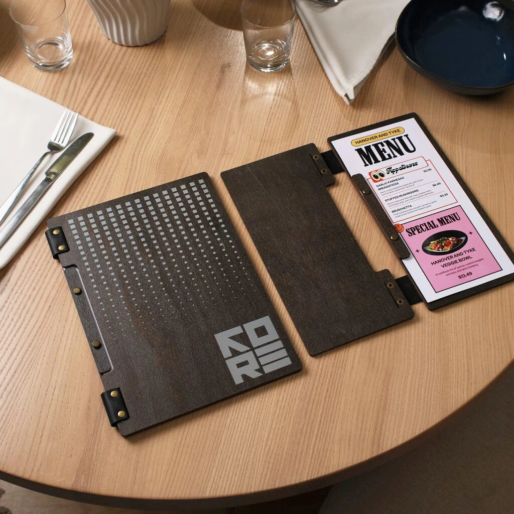 Two wenge wooden menu folders are displayed on a table next to cutlery. One folder is closed, showing its grey UV-printed geometric design, while the other is open, revealing a colorful printed menu insert with food images.